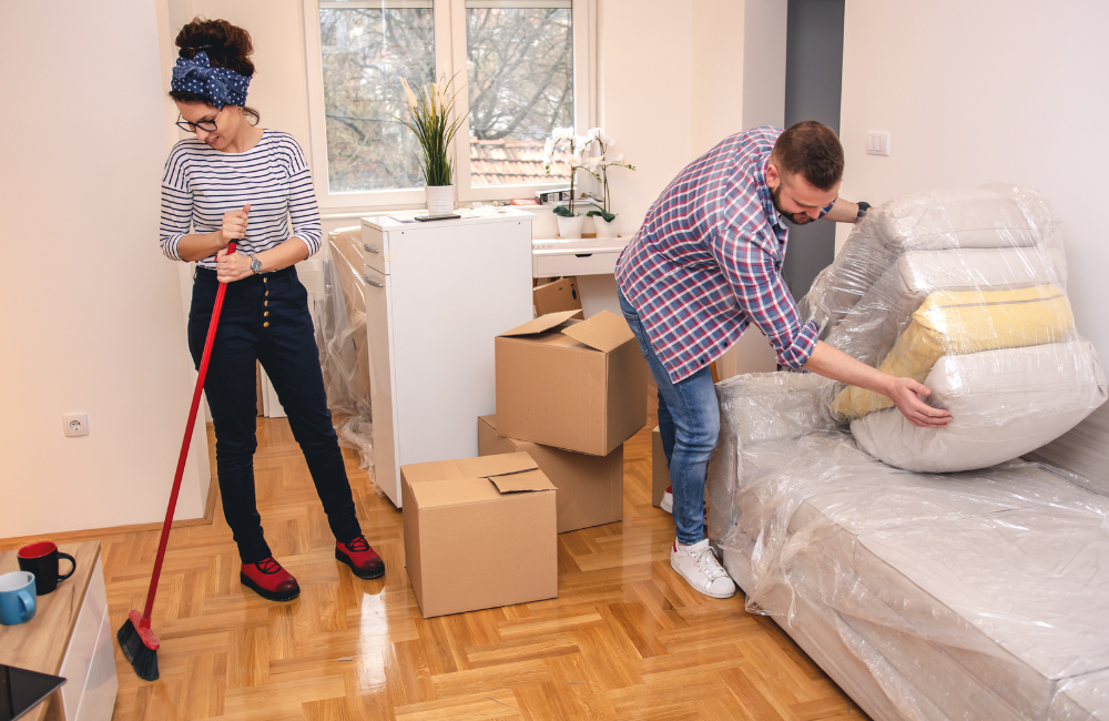 A couple in a house with all their belonging packed up ready to move. The woman is holding a broom while the man is stacking couch cushions wrapped in plastic.