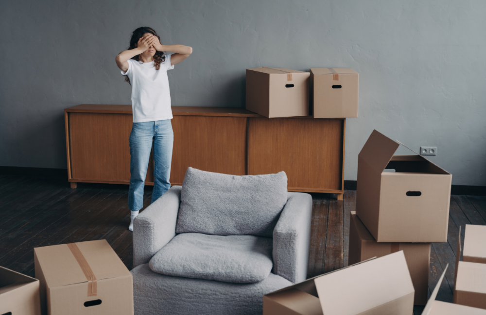 A woman standing in her lounge room with various moving boxes and furniture around her. Her hands are covering her face expressing stress.