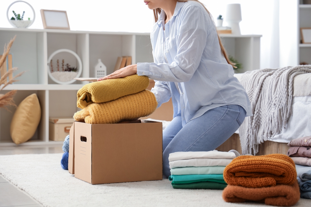 A woman kneeling next to a box and packing a pile of clothes into it.