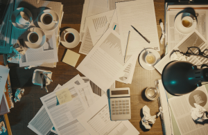 A messy desk with various office supplies, pieces of paper, and half drunk coffee cups scattered amongst work documents.