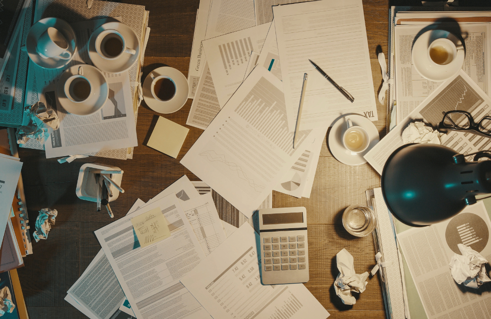 A messy desk with various office supplies, pieces of paper, and half drunk coffee cups scattered amongst work documents.