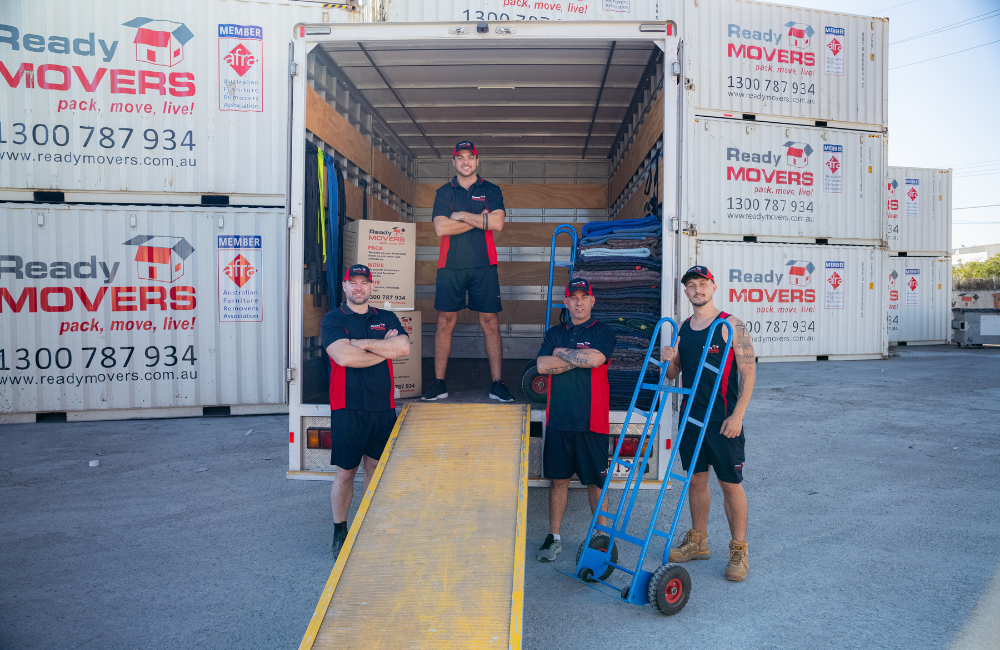 A Ready Movers team of removalists standing at the ready at the end of an open truck in a shipping container yard filled with Ready Movers shipping containers.