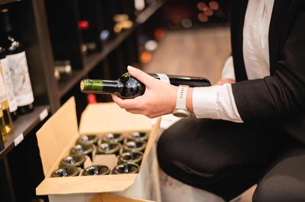 A person holding a wine bottle and placing it into a box already filled with wine. 