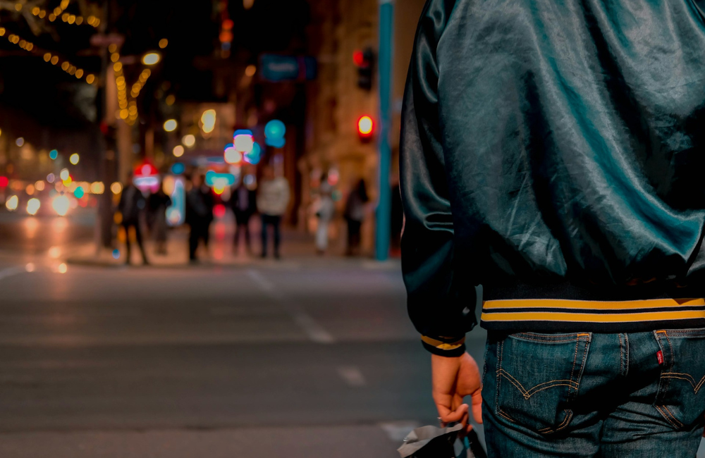 A person standing on a city street corner wearing a bomber jacket and holding a backpack.