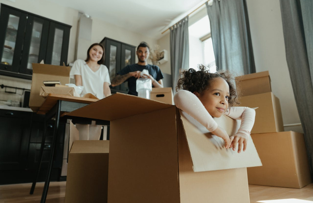 A family surrounded by boxes in their kitchen. Their daughter is sitting in one of the boxes while the mother looks on smiling.