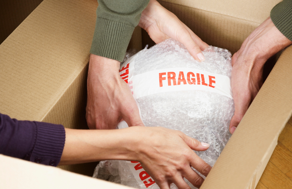 Two peoples' hands placing an object wrapped in bubble wrap and fragile tape into a box.