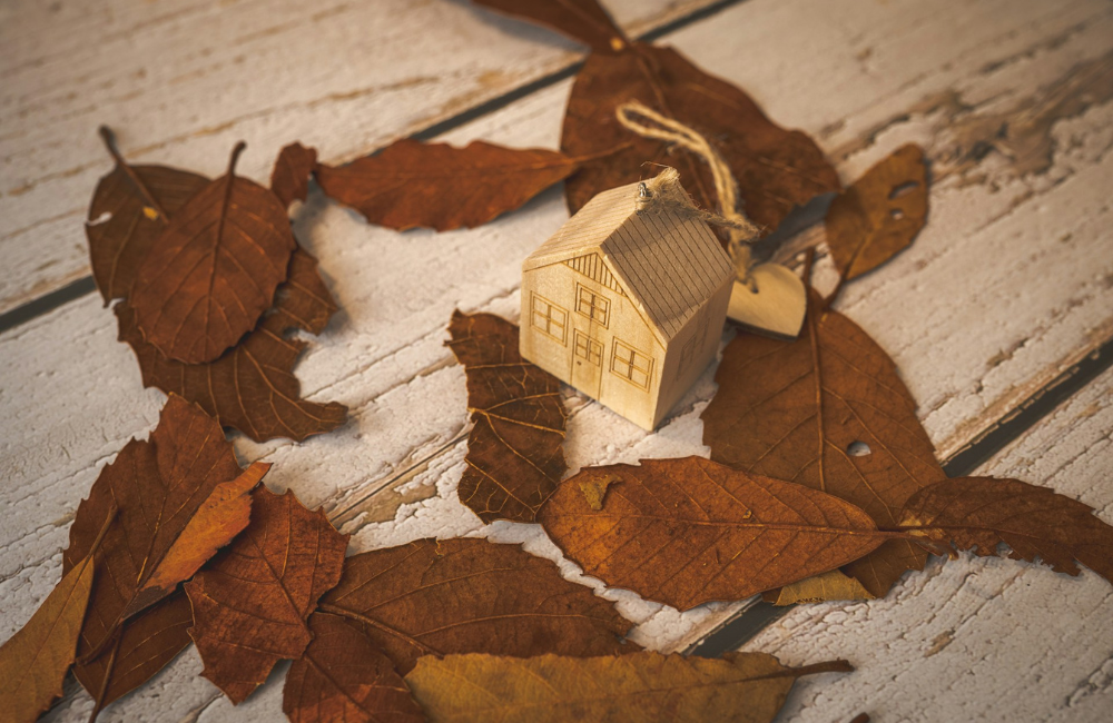 A little wooden house on a wooden table surround by brown Autumn leaves. 