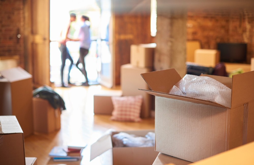 A couple standing outside talking out of focus in the background with their house filled with moving boxes in the foreground.