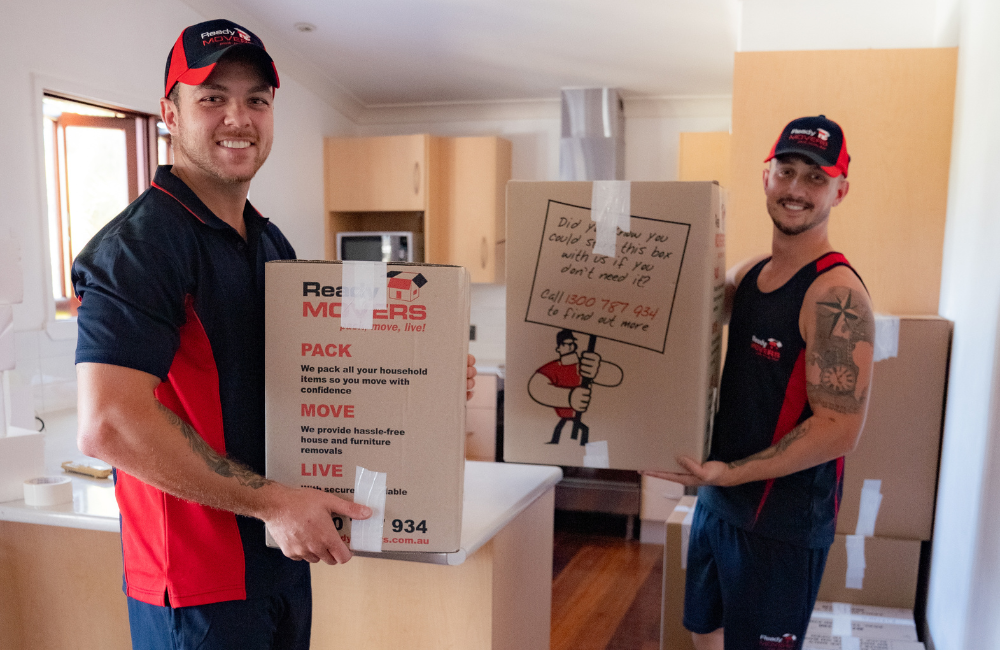 Two Ready Movers removalists standing in a person's kitchen smiling and carrying boxes.
